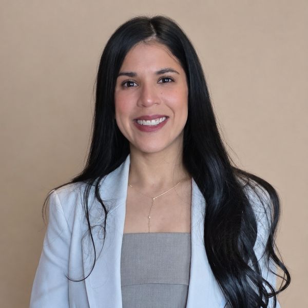 Portrait of a smiling woman with long black hair wearing a light blue blazer against a beige background.
